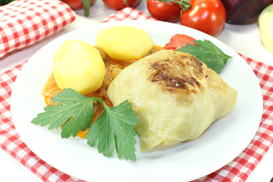 Krautroulade auf Letscho mit Salzkartoffeln, dekoriert mit Petersilieblättern auf einem weißen Teller, auf einem rot-weiß karierten Deckerl.